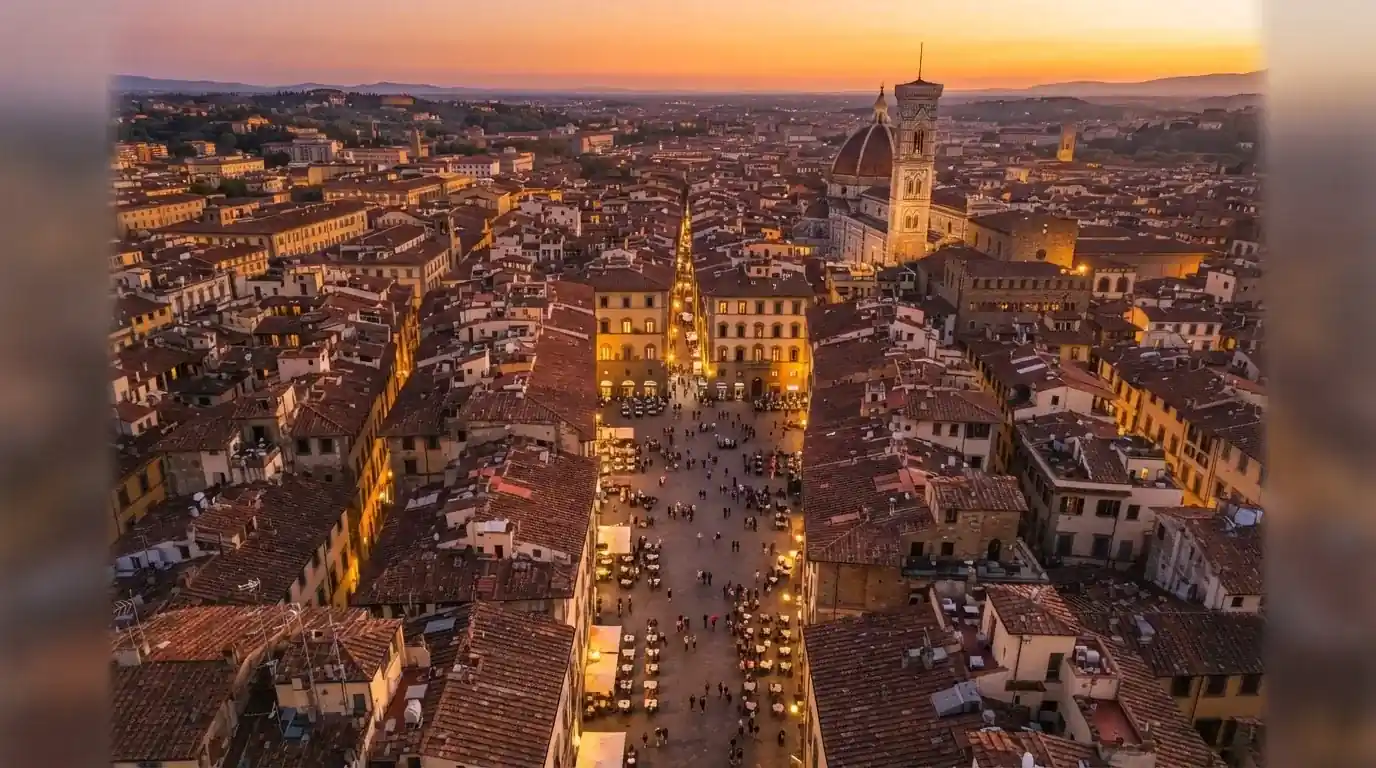Vista dall'alto di una città italiana al tramonto, con luci che si accendono nelle finestre degli edifici lungo una via trafficata.
